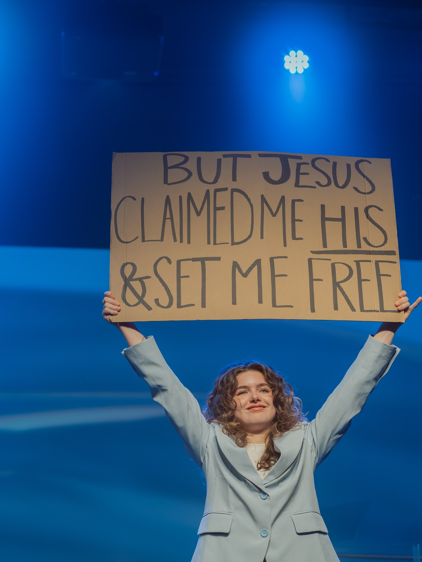 A woman on stage holding a cardboard sign that reads "BUT JESUS CLAIMED ME HIS & SET ME FREE" under blue worship lighting