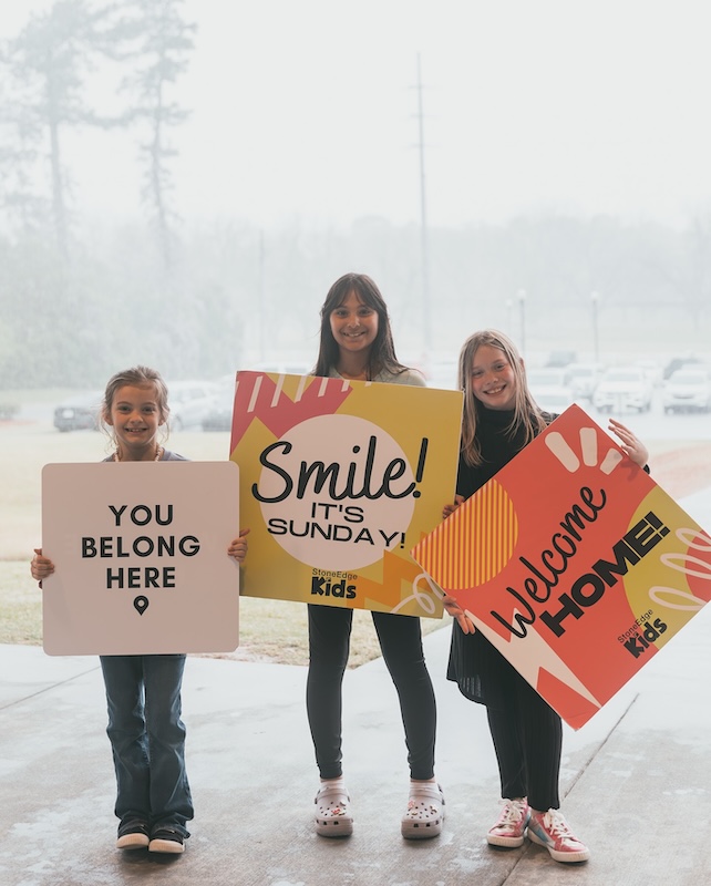 Three children hold colourful Stone Edge Kids welcome signs outside the church building in Macon, Georgia